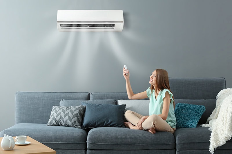 Why Are They Called Ductless Mini Splits? Photo of a woman sitting on her couch adjusting her ductless AC Unit.